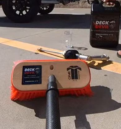 Deck Devil cleaning brush with a visible brand logo on a concrete surface, with a bottle of Ultra Wash cleaning solution in the background.